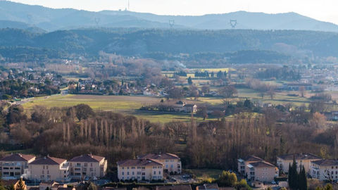 Vue panoramique d’une vallée rurale: maisons, champs et arbres au pied de collines bleutées sous une lumière douce.