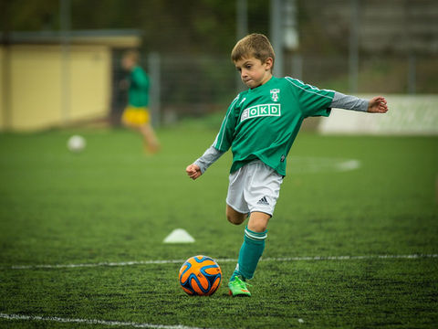 Jeune footballeur en maillot vert frappe un ballon orange sur un terrain, avec le logo OKD sur le torse.