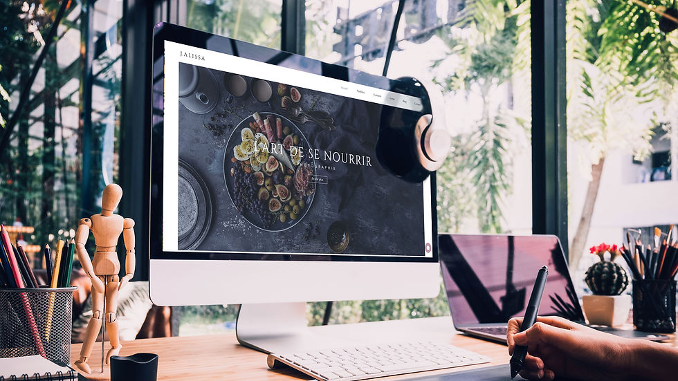 Bureau créatif avec iMac affichant L’ART DE SE NOURRIR, main dessinant sur tablette, palettes et lumière de plantes.