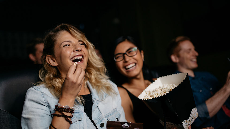 Deux femmes rient aux éclats dans une salle de cinéma sombre, l’une mange du pop-corn, gobelets et sachets visibles.