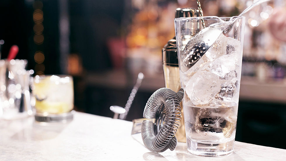 Verre à cocktail rempli de glaçons avec passoire, sur un comptoir de bar, fond flou aux lumières chaudes.