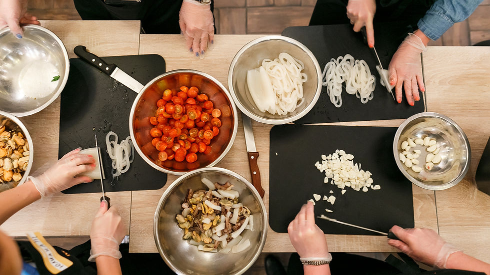 Des personnes découpent des légumes sur des planches noires. Bols en métal avec tomates cerises, ail, champignons. Ambiance culinaire animée.
