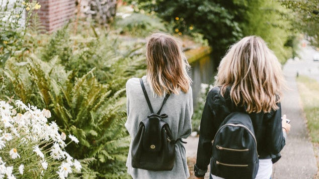 Deux femmes avec sacs à dos marchent sur un chemin de jardin près d’une maison et de fleurs blanches.