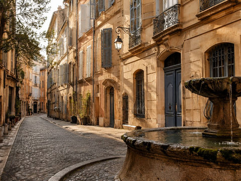 Rue pavée paisible dans une vieille ville, fontaine au premier plan, façades beige aux volets bleus, sans passants.
