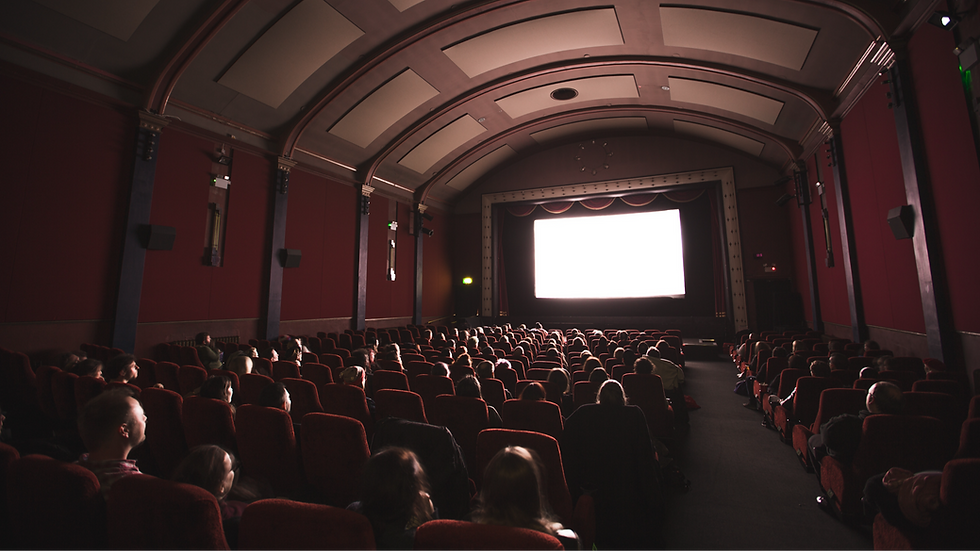 Salle de cinéma sombre avec public assis sur sièges rouges, grand écran blanc allumé, ambiance attentive.