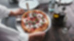Une main sert une pizza garnie de basilic, fromage, et tomates séchées sur une table en bois. Huiles, verres, et assiettes en arrière-plan.