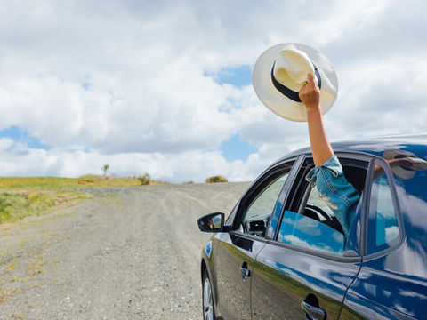 Bras hors d’une voiture noire, tenant un chapeau blanc, sur une route de gravier sous un ciel nuageux.
