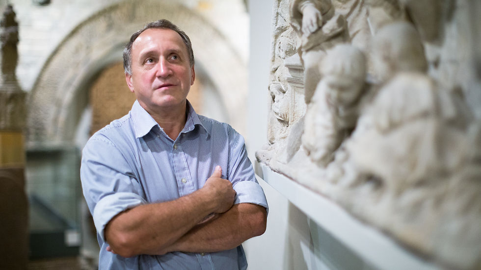 Homme en chemise bleue dans un musée, bras croisés, contemplant des reliefs sculptés en pierre claire.