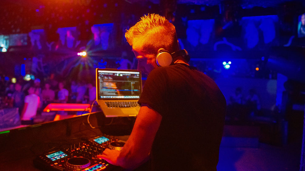 DJ blond au casque mixe sur un ordinateur portable et une console, dans un club sombre aux lumières bleues et orange.