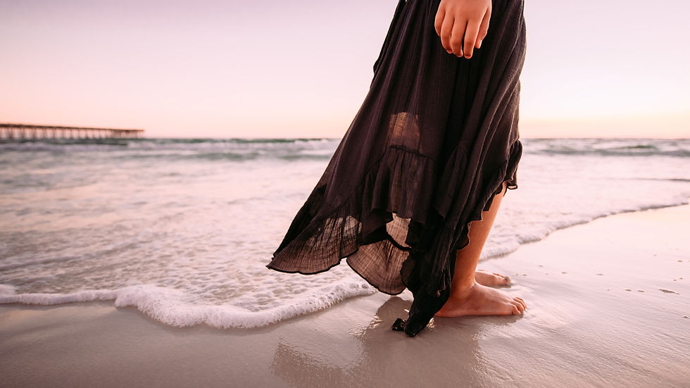 Femme pieds nus en robe noire sur la plage au coucher du soleil, vagues douces et ponton au loin.