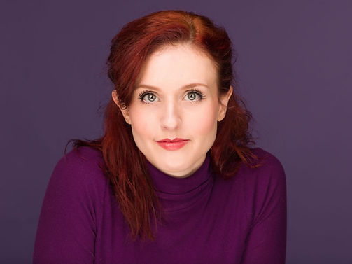 Headshot of Hadley - a red haired woman wearing a purple turtleneck, set against a solid purple backdrop. 