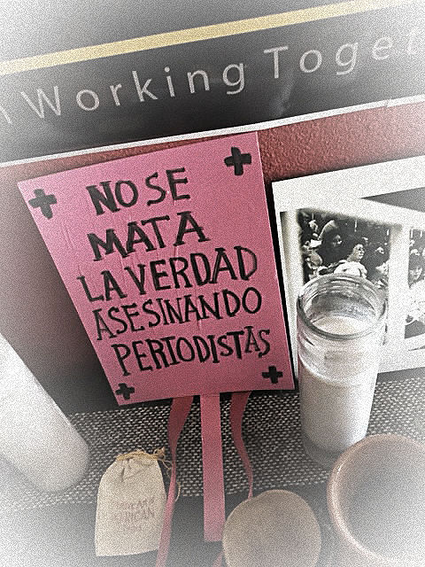 Pink sign with "No se mata la verdad asesinando periodistas" text, candle, framed photo, and cups on a table, evoking a somber mood.