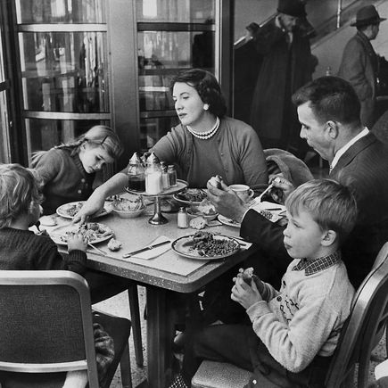 Family dining at a restaurant table, sharing food and conversation. People in coats visible through glass. Warm, lively atmosphere.