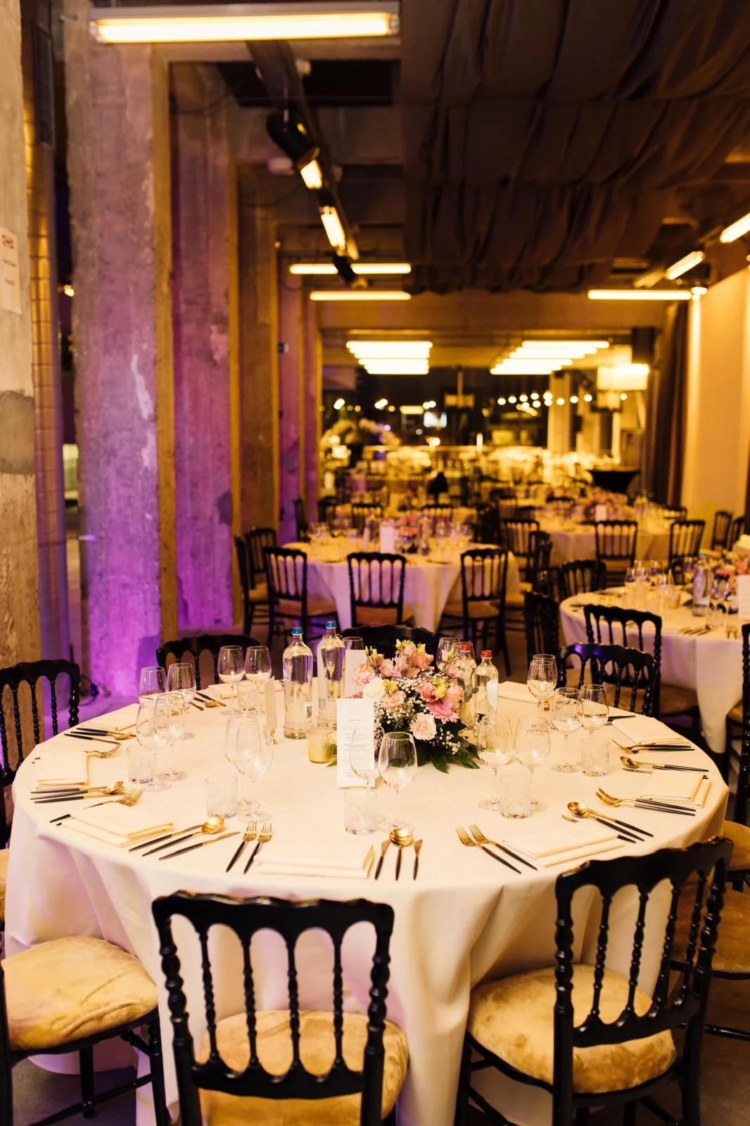 Table ronde dressée pour un événement, avec nappage blanc, verres, bouteilles d'eau et un centre de table fleuri. L'éclairage d'ambiance est doux et coloré.