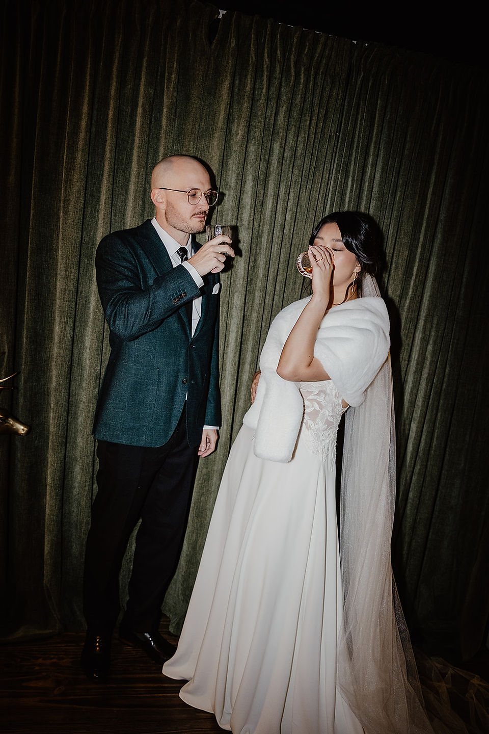 The bride and groom share a whiskey in front of a green velvet curtain inside their Fremantle Airbnb