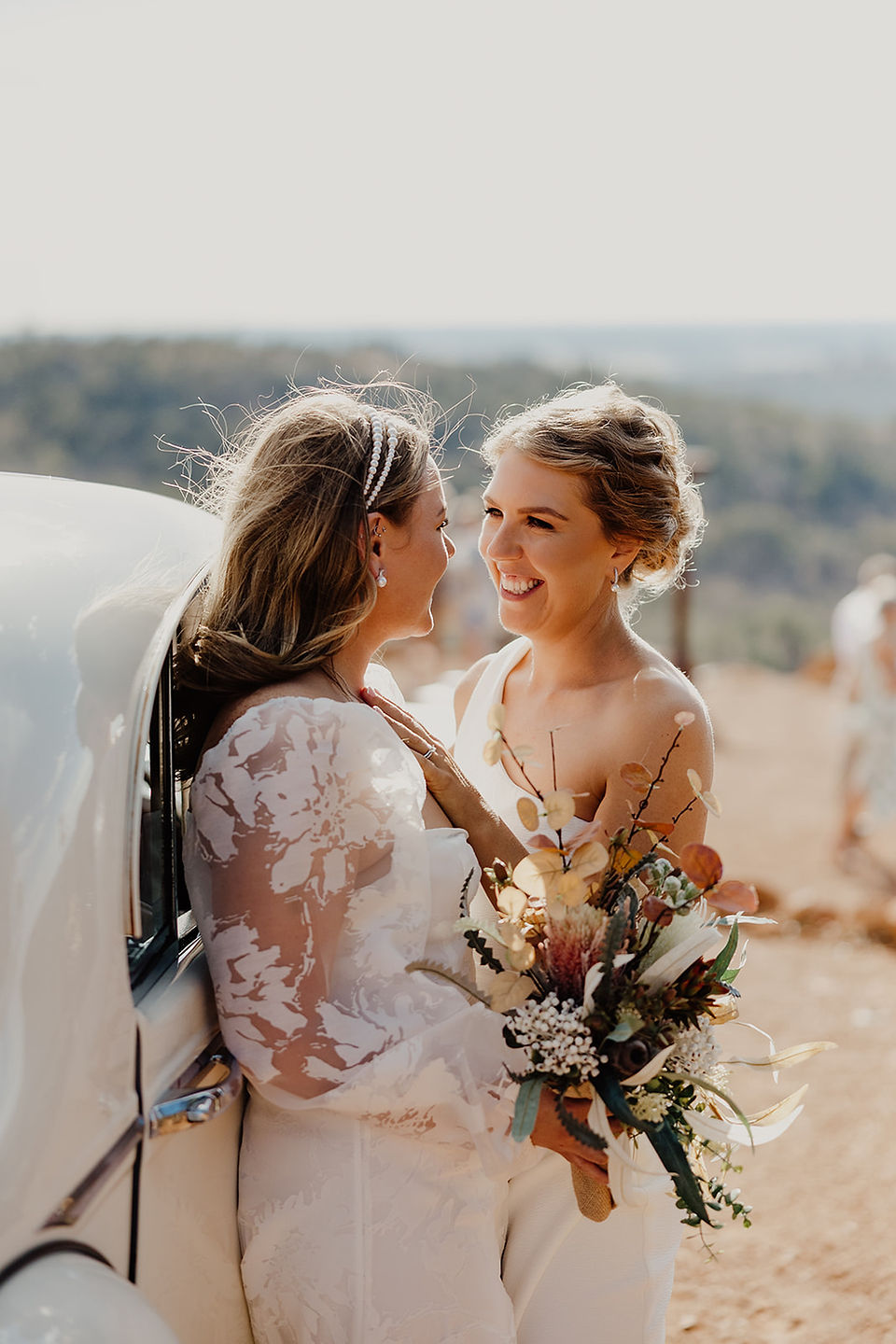 Two brides wear white wedding dresses and hold bouquets as they lean against a classic Rolls Royce at their wedding ceremony at Anderson Point in the Perth HIlls