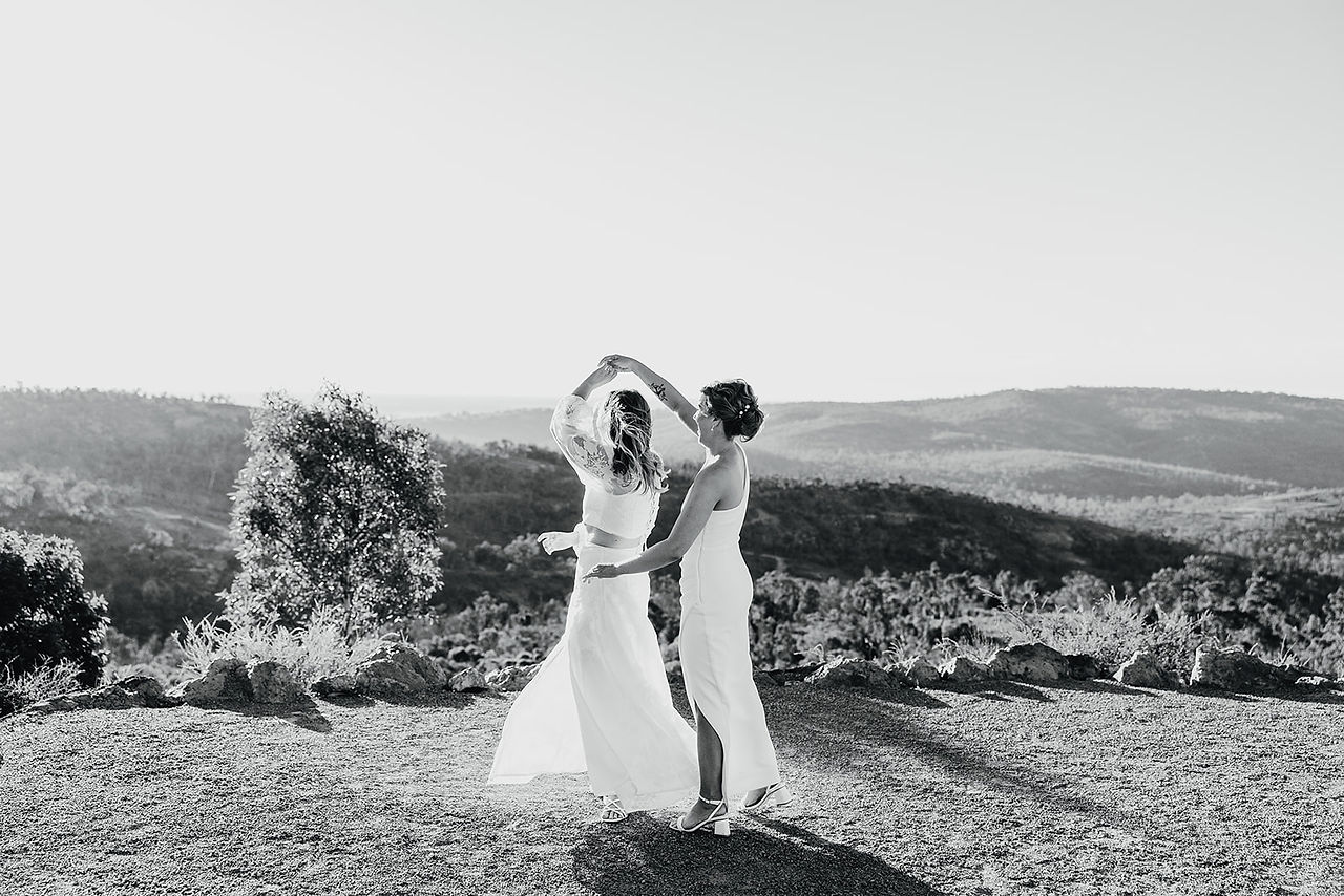 Two brides smile at each other as they dance with the view of the national park behind them. They are at their wedding ceremony venue at Anderson Point, surrounded by the Australian bush in the Perth HIlls.
