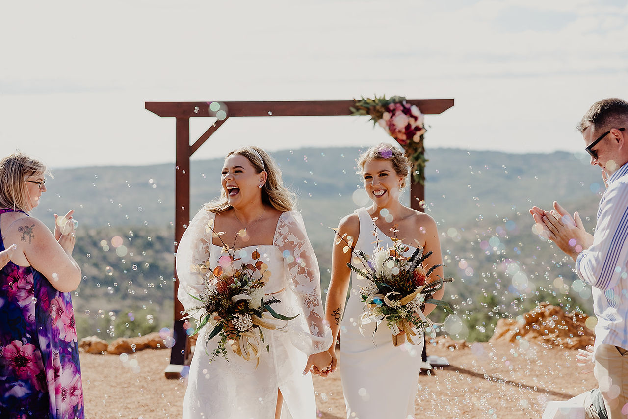 Two brides hold hands as they exit their wedding ceremony at Anderson Point in the Perth Hills. They stand in front of a wooden arbor adorned with flowers and bubbles are blowing in front of them. 