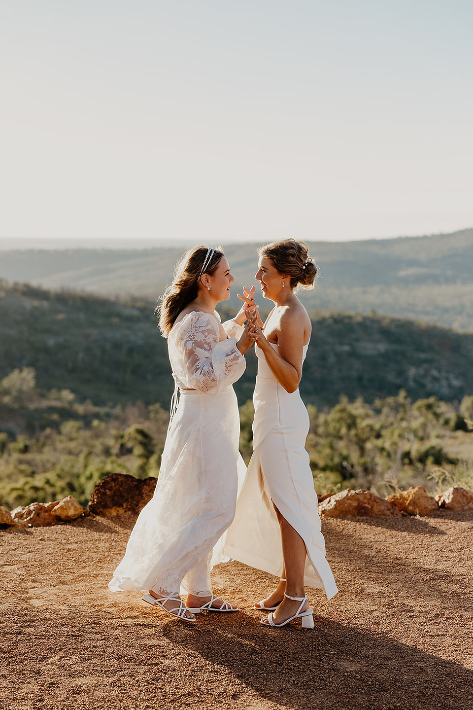 Two brides wearing white dresses hold hands and dance slowly on the hillside. They are at their wedding ceremony venue at Anderson Point, surrounded by the Australian bush in the Perth HIlls.