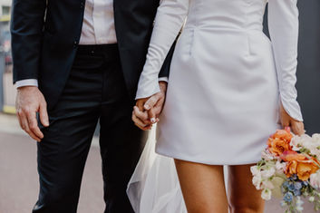 The legs of a bride and groom. The bride wears a fitted short wedding dress and holds a small posy of flowers.