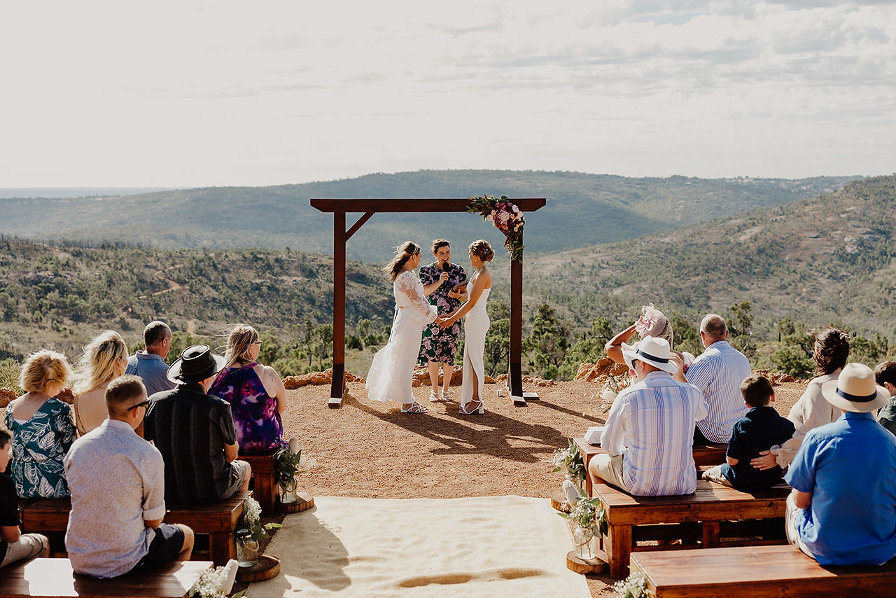 Two brides hold hands during their wedding ceremony on the hillside ceremony space at Anderson Point with the national park as the natural backdrop