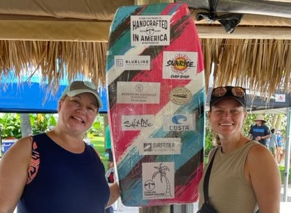 A couple of women standing on both sides of a surfboard used to promote the event.