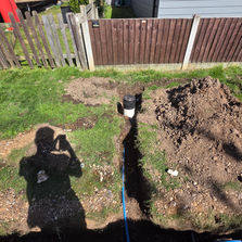 A long dirt trench with a blue pipe running along a black barn building with a red container structure on the side.
