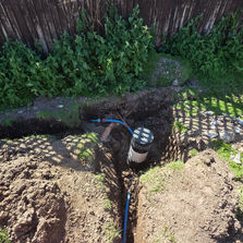 A narrow trench with a blue pipe running down the center of a grassy path, with an excavator facing forward at the top of the trench.