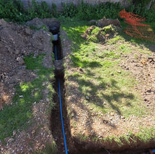 A long dirt trench with a blue pipe running along a black barn building with a red container structure on the side.