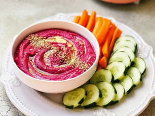 Plato blanco con rodajas de pepino, bastones de zanahoria y un bowl blanco con hummus de remolacha