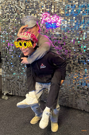 Zwei Personen tragen sich, "Party Time" und "BOOM!" auf glitzerndem Hintergrund.