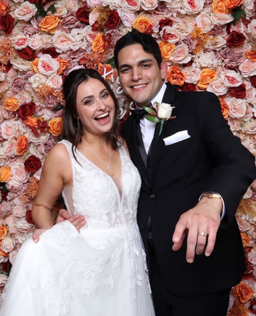 Lächelndes Brautpaar vor einer opulenten Wand aus roten und orangefarbenen Rosen.