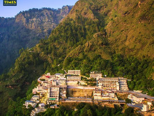 Mountain town of white buildings nestled on a green cliff, with Tour Times logo at top left and bright blue sky overhead Vaishno Devi Temple