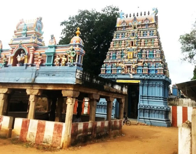 Colorful Hindu temple entrance with ornate blue-and-pastel gopuram beside a dirt path, trees, and striped walls.Thiru Kabisthalam Temple