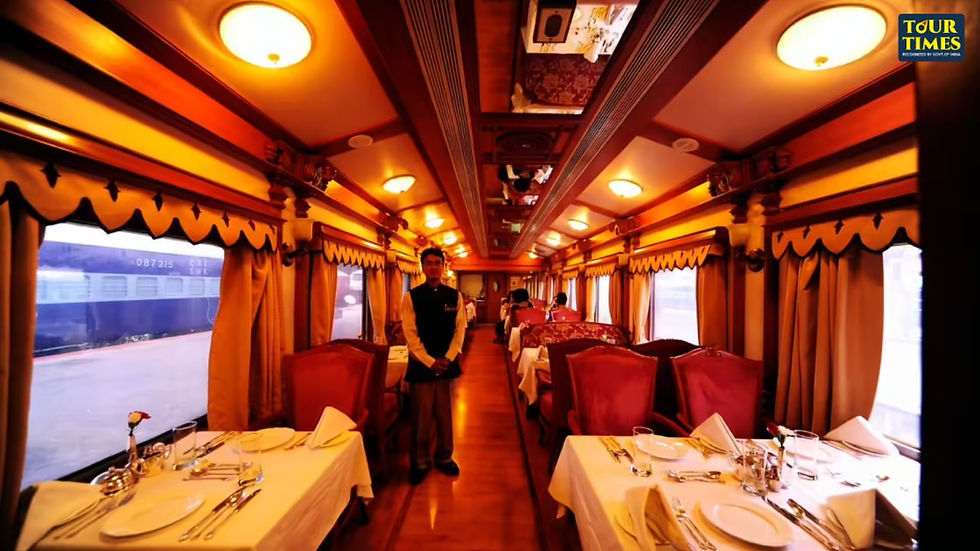 Luxurious train dining car with elegant decor, red chairs, and white tablecloths. A staff member stands in the aisle. Warm lighting. Karnataka luxury tour
