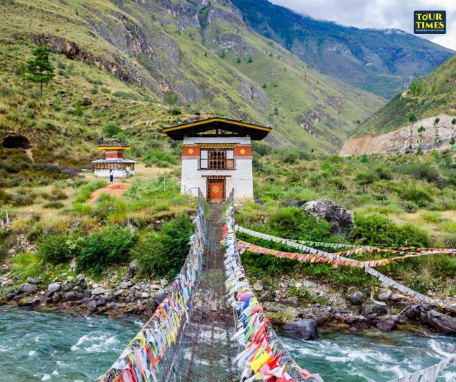 Suspension bridge with colorful prayer flags over a river, leading to traditional Bhutanese buildings in a lush mountain landscape. Tour Times logo. Famous Place in Bhutan