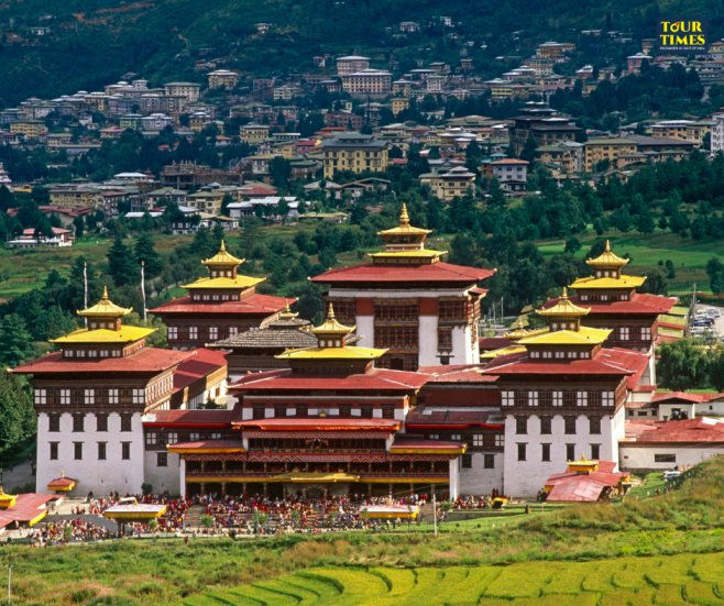 Majestic Bhutanese temple with red and gold roofs, surrounded by lush greenery and a distant town. Bright, celebratory mood. Tour Times text. Famous Place in Bhutan