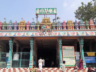 Colorful Hindu temple entrance with ornate statues and signs, a few people standing and walking below, under a clear sky. Thiruthangal Temple