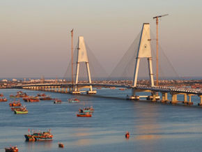 Bridge spans a river with several colorful boats; cityscape in the background. Soft twilight colors, "Tour Times" text in the corner.Bet Dwarka Temple