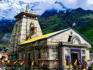 Stone Himalayan temple with colorful flags and a few visitors, snowy mountains and clouds behind; TOUR TIMES watermark. Kedarnath Jyotirlinga Temple