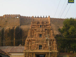 Ornate temple gopuram before a fort wall on a rocky hill, with trees and power lines; Tour Times logo in corner. Thirumayam Perumal Temple