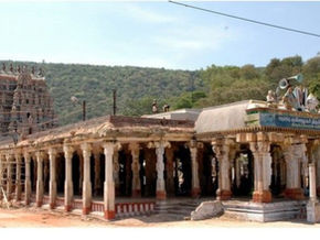Ancient South Indian temple with carved columns and gopuram against green hills, plus a Tour Times watermark.