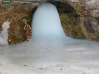 A large ice stalagmite inside a rocky cave. A trident adorned with cloth and bells leans against it. Text: Tour Times at the top left.Amarnath Cave Temple
