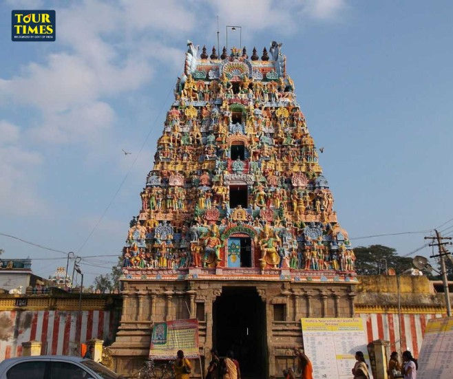 Colorful Hindu temple gopuram with statues against blue sky, people below, and a Tour Times logo in the corner. Thiru Thanjai Mamani Koil