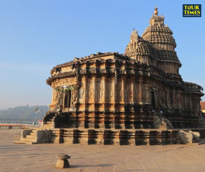 Ancient stone temple with carved tower under clear blue sky, Tour Times logo in corner. Karnataka Temple Tour