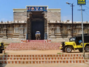 Colorful Hindu temple gopuram rises against blue sky, framed by palm leaves and a concrete beam; Tour Times logo at top right.