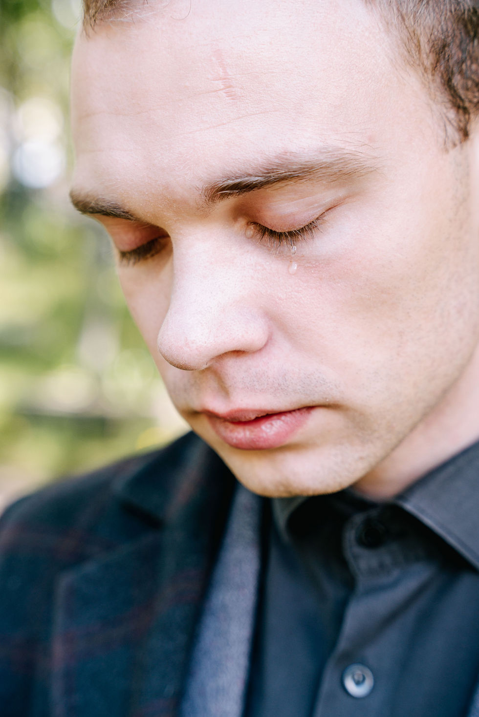 Close-up of a young man with eyes closed and a single tear rolling down his cheek, conveying vulnerability and emotional depth in a softly blurred natural setting.