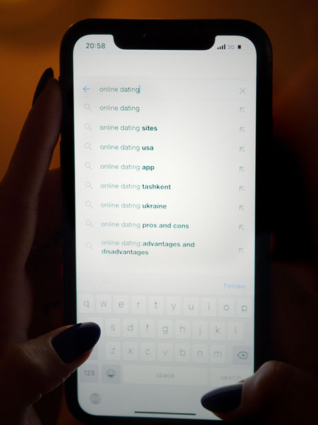 A person's hands hold a smartphone in a dimly lit room, displaying search suggestions for "online dating" including "pros and cons" and "advantages and disadvantages," illustrating the analytical mindset behind app-based mate selection.