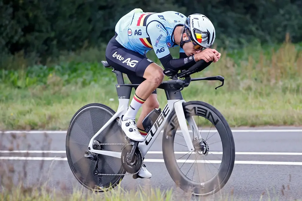 Cyclist in a blue jersey rides a sleek white bike on a road, leaning forward intensely. Green foliage in the background; visible brand logos.
