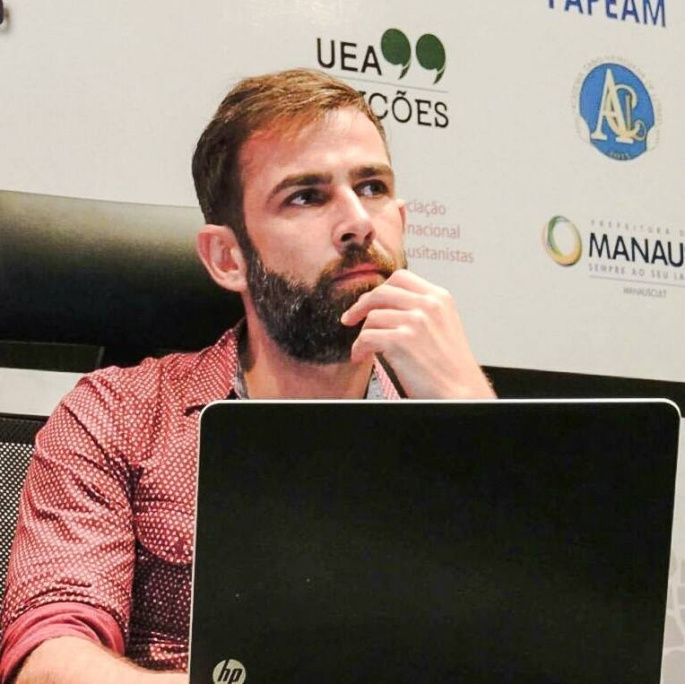 Foto de Rodrigo Borba: homem branco, loiro, de cabelo curto e branca curta e grisalha. Usa uma camisa rosa, com pontinhos brancos. Em primeiro fundo na foto, a parte detrás da tela de um notebook, que está em frente ao convidado. Ao fundo, um painel com alguns logotipos de eventos e associações científicas.